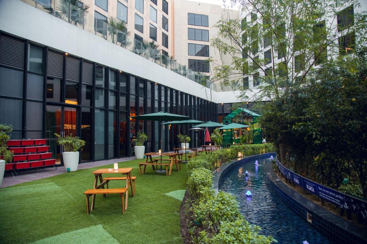 Novotel New Delhi Aerocity Екстериор снимка The photo shows a modern outdoor space that appears to be part of a hotel or a recreational area. There are several wooden tables with benches arranged on a green lawn, suggesting a place for guests to relax or dine outdoors. In the background, there