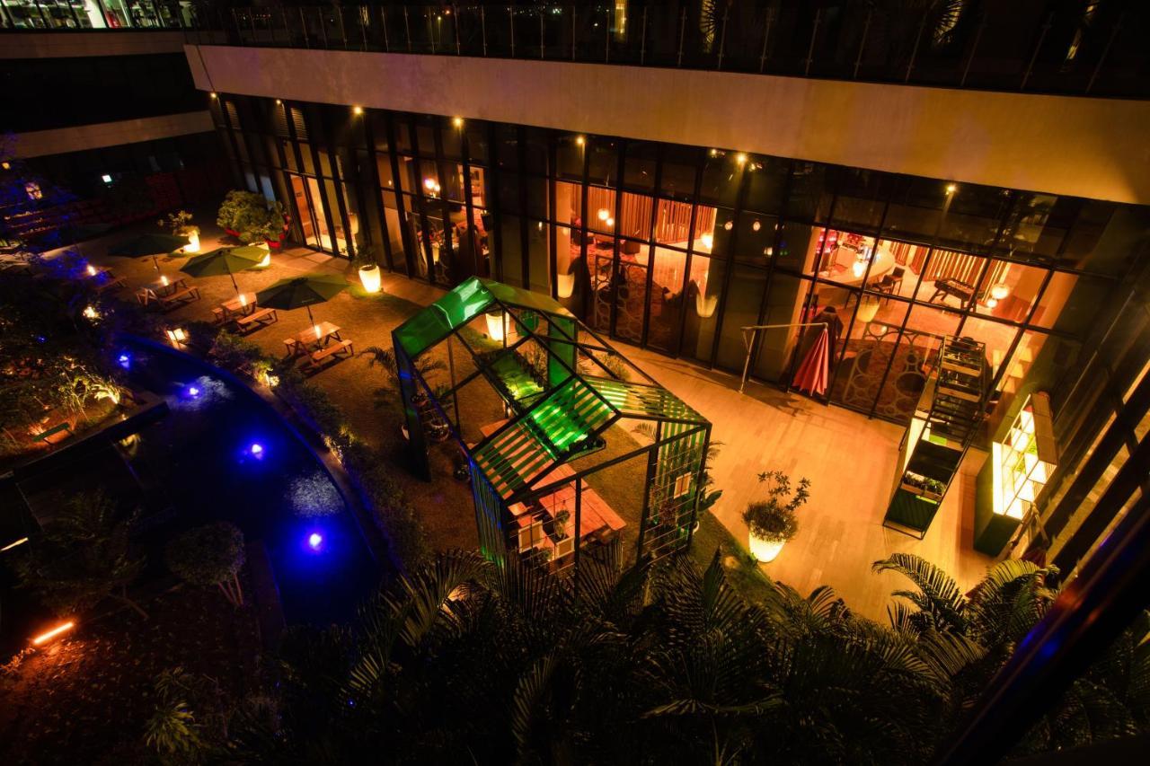 Novotel New Delhi Aerocity Екстериор снимка The photo shows an outdoor area of a restaurant or café at night. The setting features a well-lit space with various seating arrangements, surrounded by greenery. In the center, there is a striking structure, possibly a gazebo, that is illuminated wi