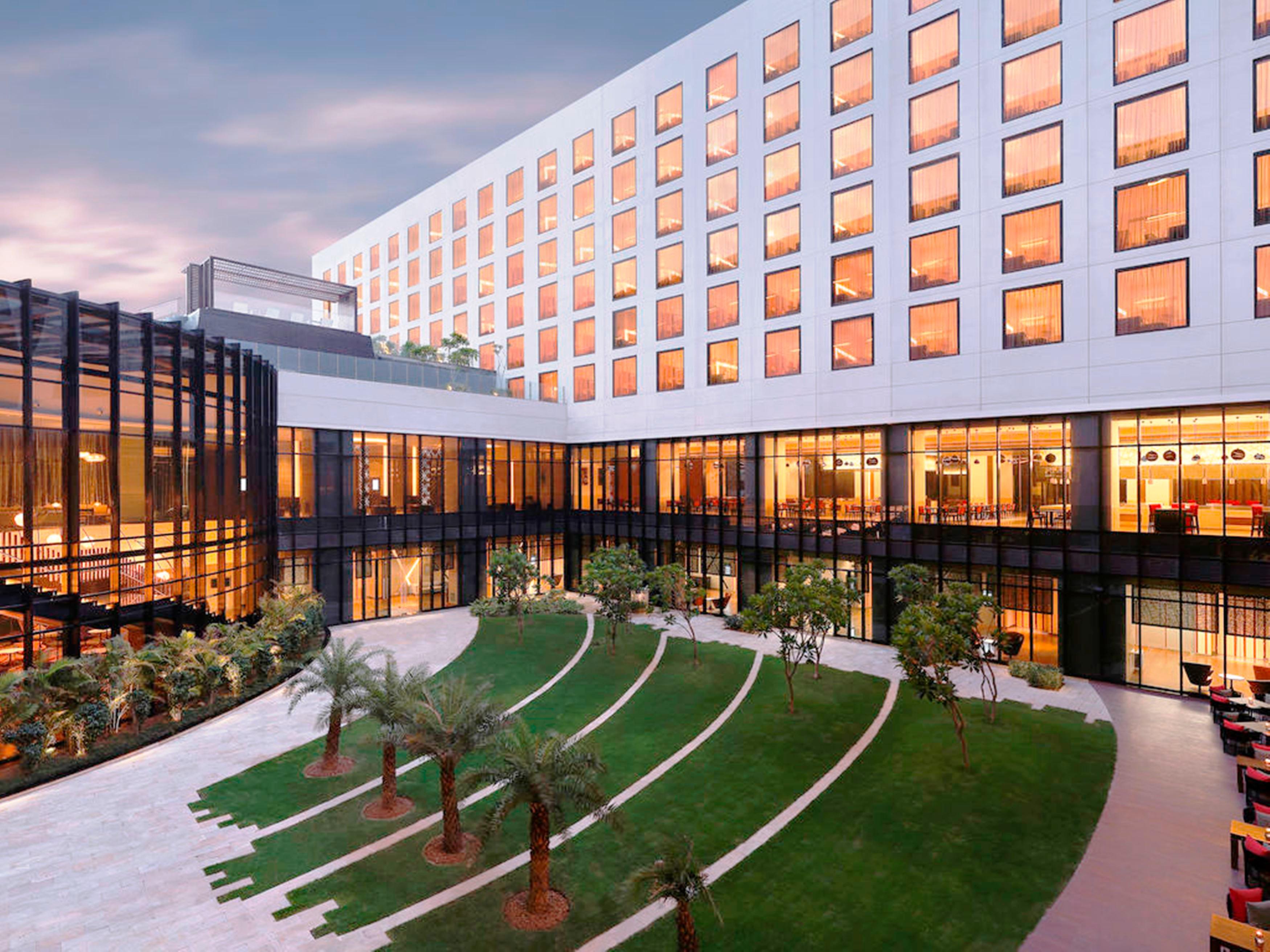 Novotel New Delhi Aerocity Екстериор снимка The photo shows a modern hotel or building complex with a sleek architectural design. In the foreground, there is a well-maintained green lawn featuring landscaped areas and small trees. The building has large glass windows that reflect the sky, and 