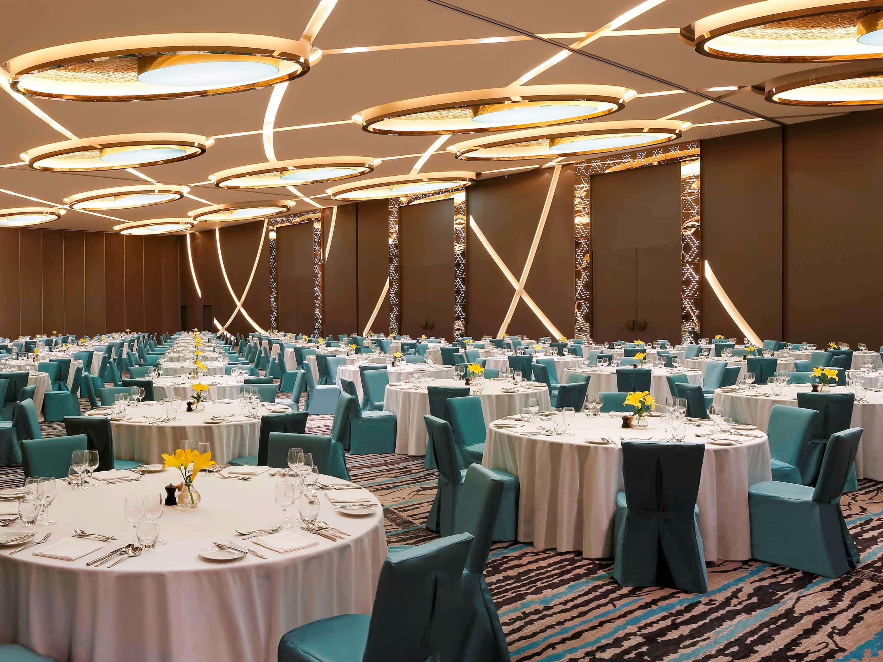 Novotel New Delhi Aerocity Екстериор снимка The photo depicts a spacious banquet hall set up for an event. There are multiple round tables covered with white tablecloths, each set with dining utensils, plates, and small vases containing yellow flowers. The chairs are upholstered in a teal fabr