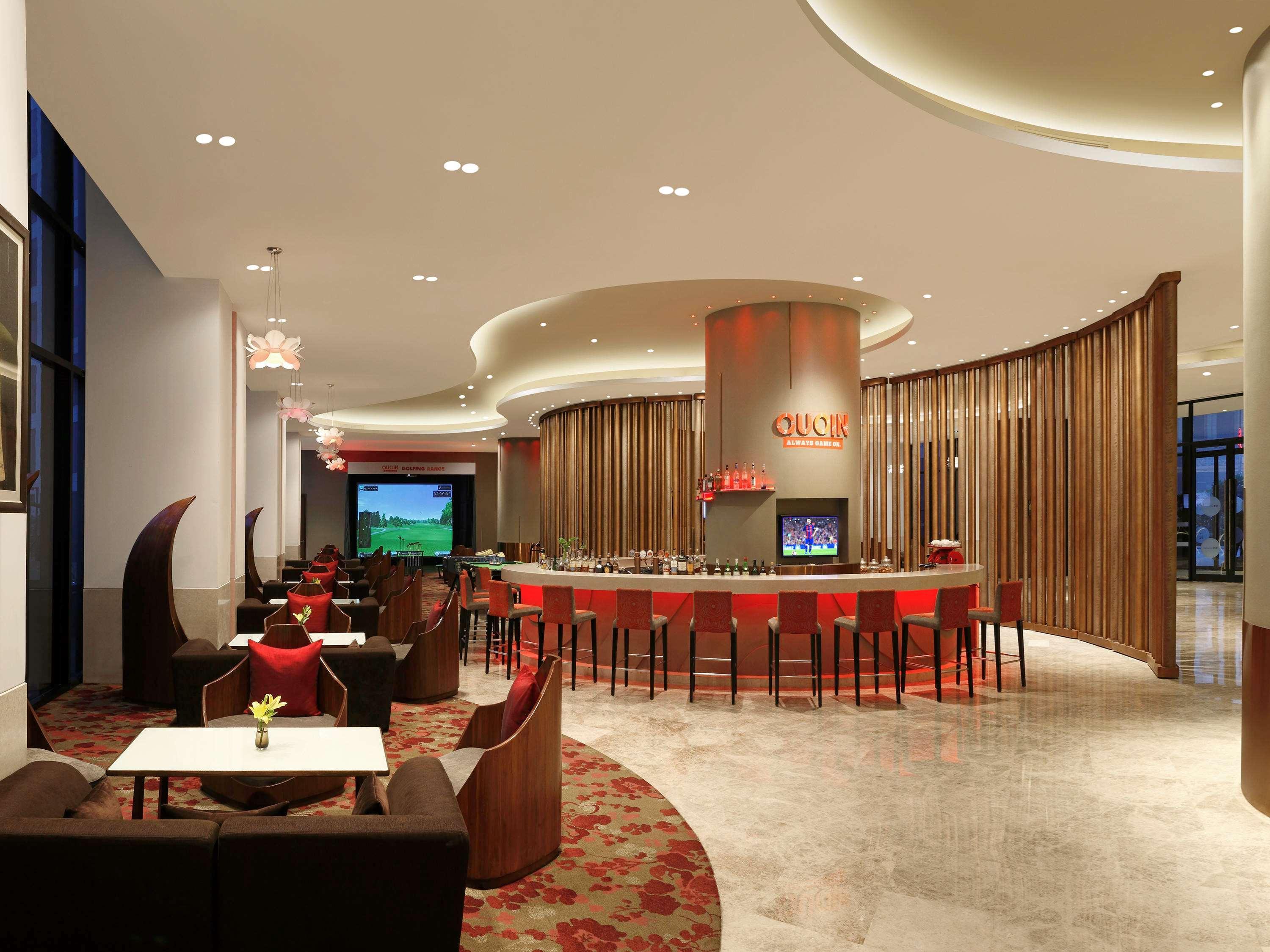 Novotel New Delhi Aerocity Екстериор снимка The photo shows a modern and stylish interior of a lounge or bar. The space features a curved bar in the center, surrounded by seating arrangements that include dark-colored couches and chairs. The design elements include a mix of smooth surfaces and