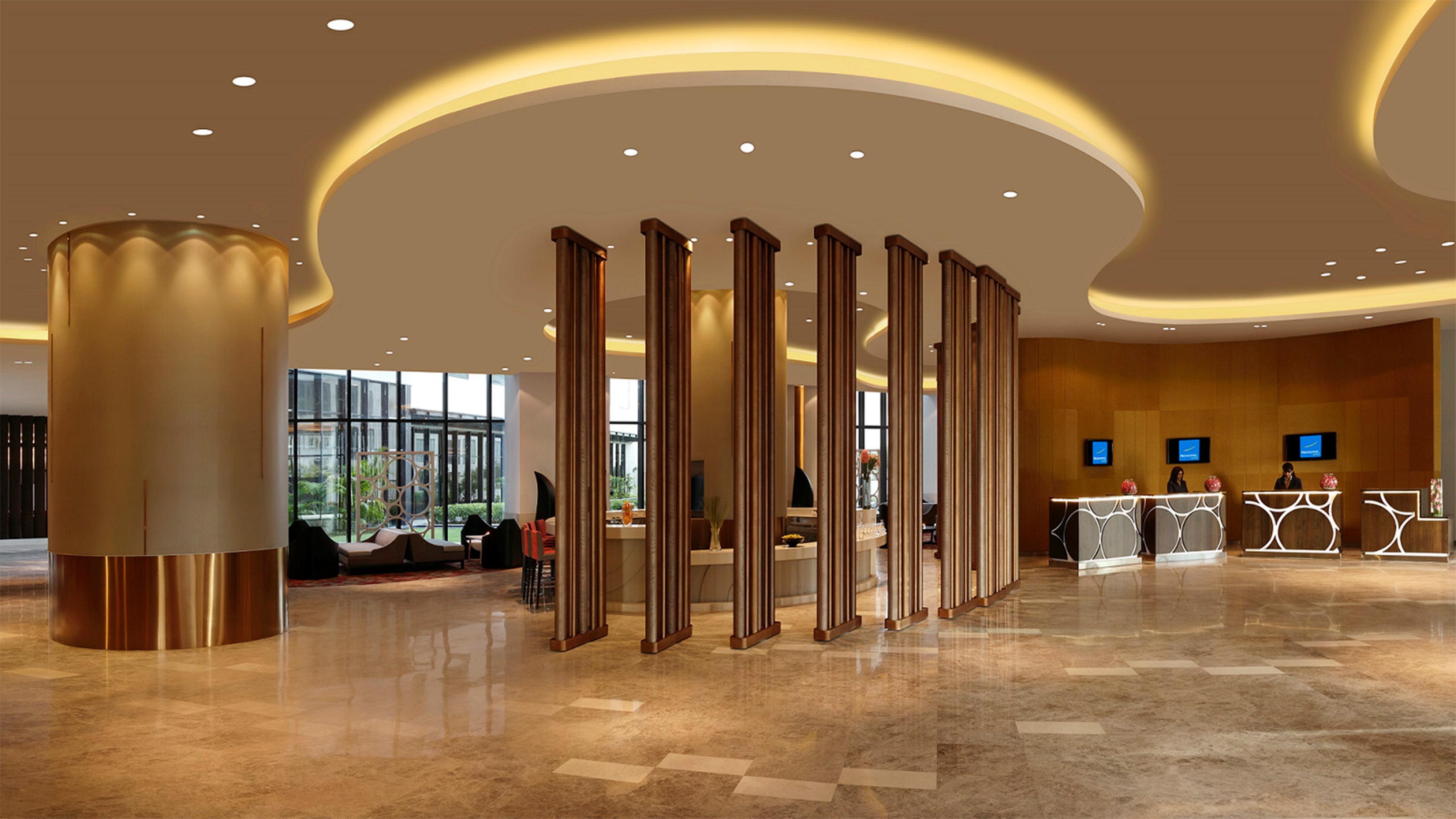 Novotel New Delhi Aerocity Екстериор снимка The photo shows a modern hotel lobby with a spacious and elegant design. The ceiling features a smooth, curved shape with ambient lighting creating a warm atmosphere. In the center, there are vertical wooden elements that add a sense of structure and