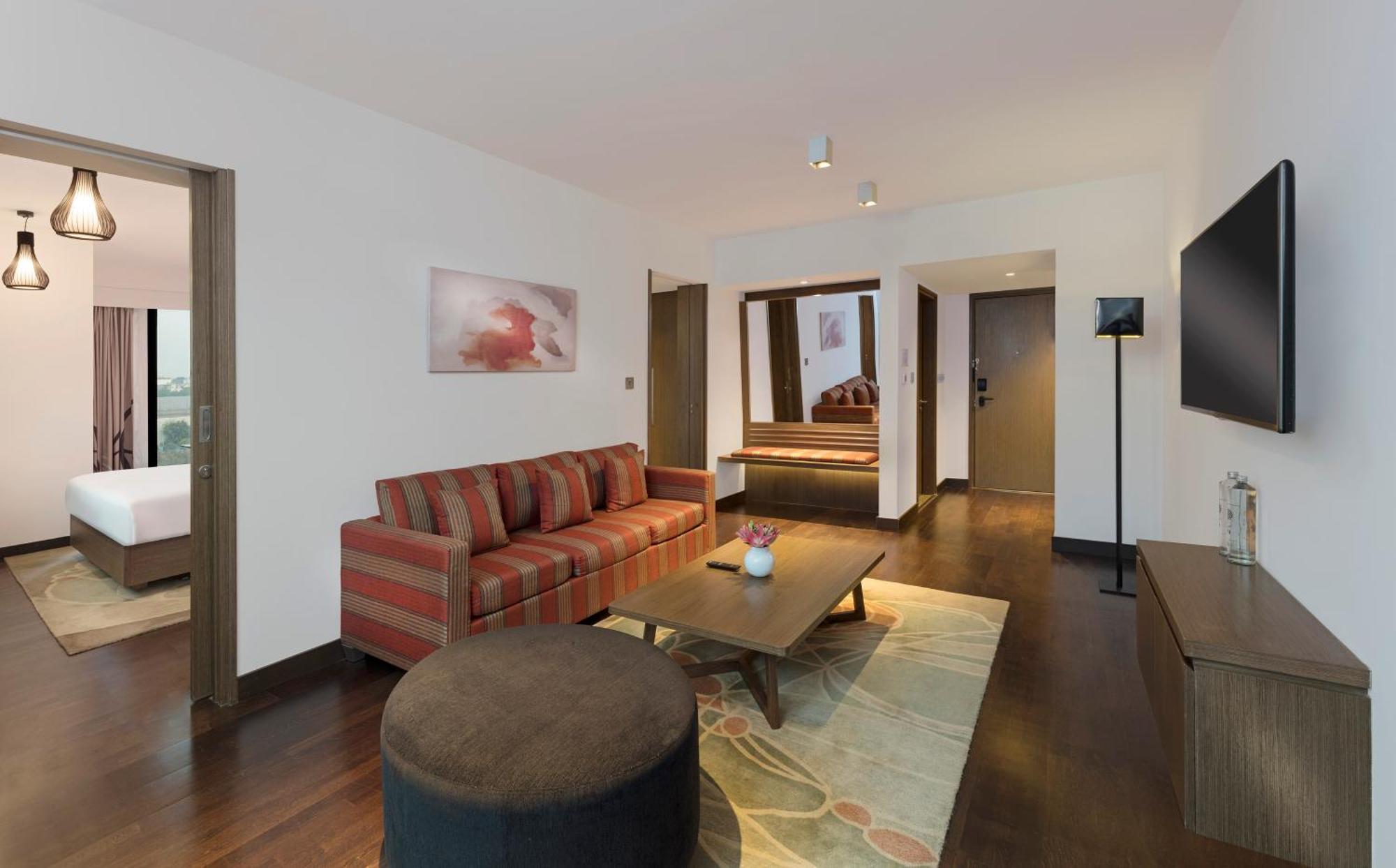 Novotel New Delhi Aerocity Екстериор снимка The photo shows a modern hotel suite or living area. It features a striped sofa with cushions, a round coffee table, and a decorative rug on a wooden floor. There is a flat-screen TV mounted on the wall, and a small side lamp nearby. The layout inclu