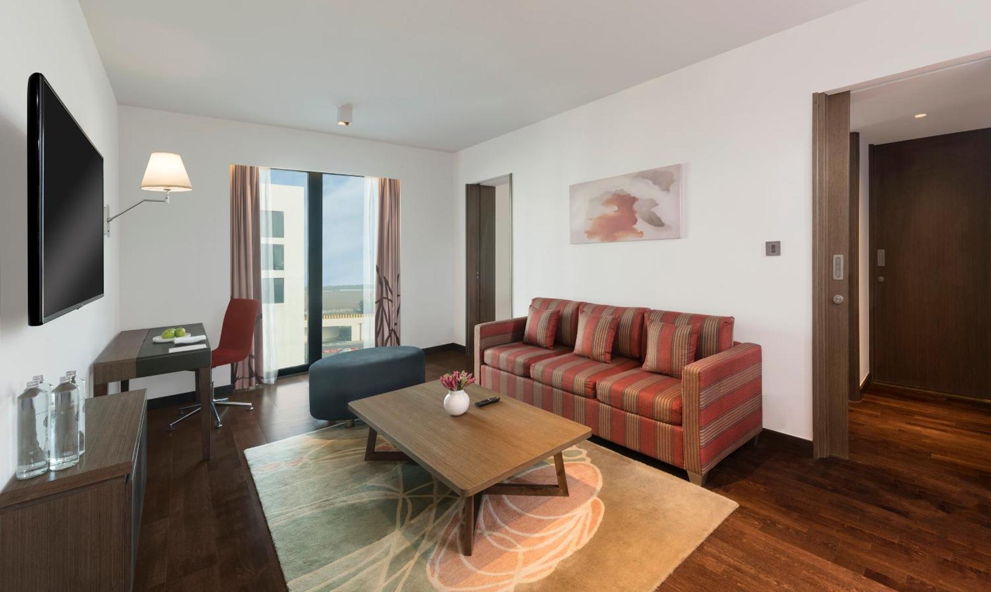 Novotel New Delhi Aerocity Екстериор снимка The photo shows a modern living room with a contemporary design. It features a striped sofa in shades of red and brown, positioned near a coffee table. The coffee table is dark in color and has a small decorative vase on it. There’s a light gray area