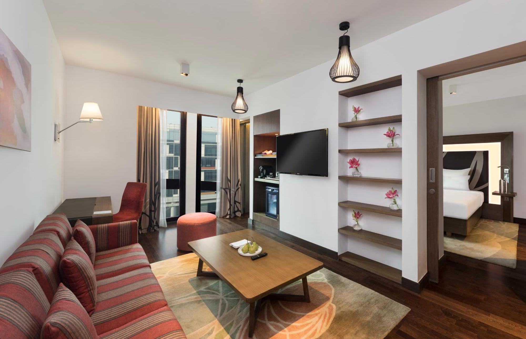 Novotel New Delhi Aerocity Екстериор снимка The photo shows a modern hotel room or suite with a cozy living area. There is a striped red and beige couch facing a wooden coffee table, which has some fruit placed on it. The room features large windows with curtains that allow natural light to en