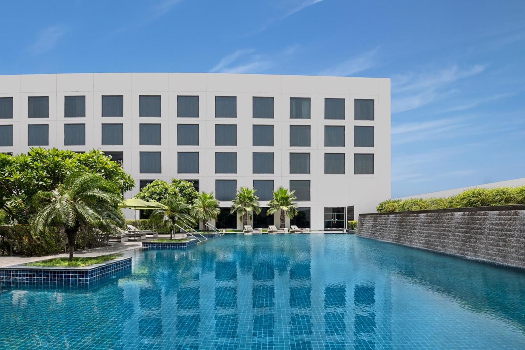 Novotel New Delhi Aerocity Екстериор снимка The photo shows a modern hotel building with a sleek, minimalist design. In front of the hotel is a large swimming pool, which reflects the blue sky and the building. Surrounding the pool are palm trees and lounging areas, creating a relaxing atmosph