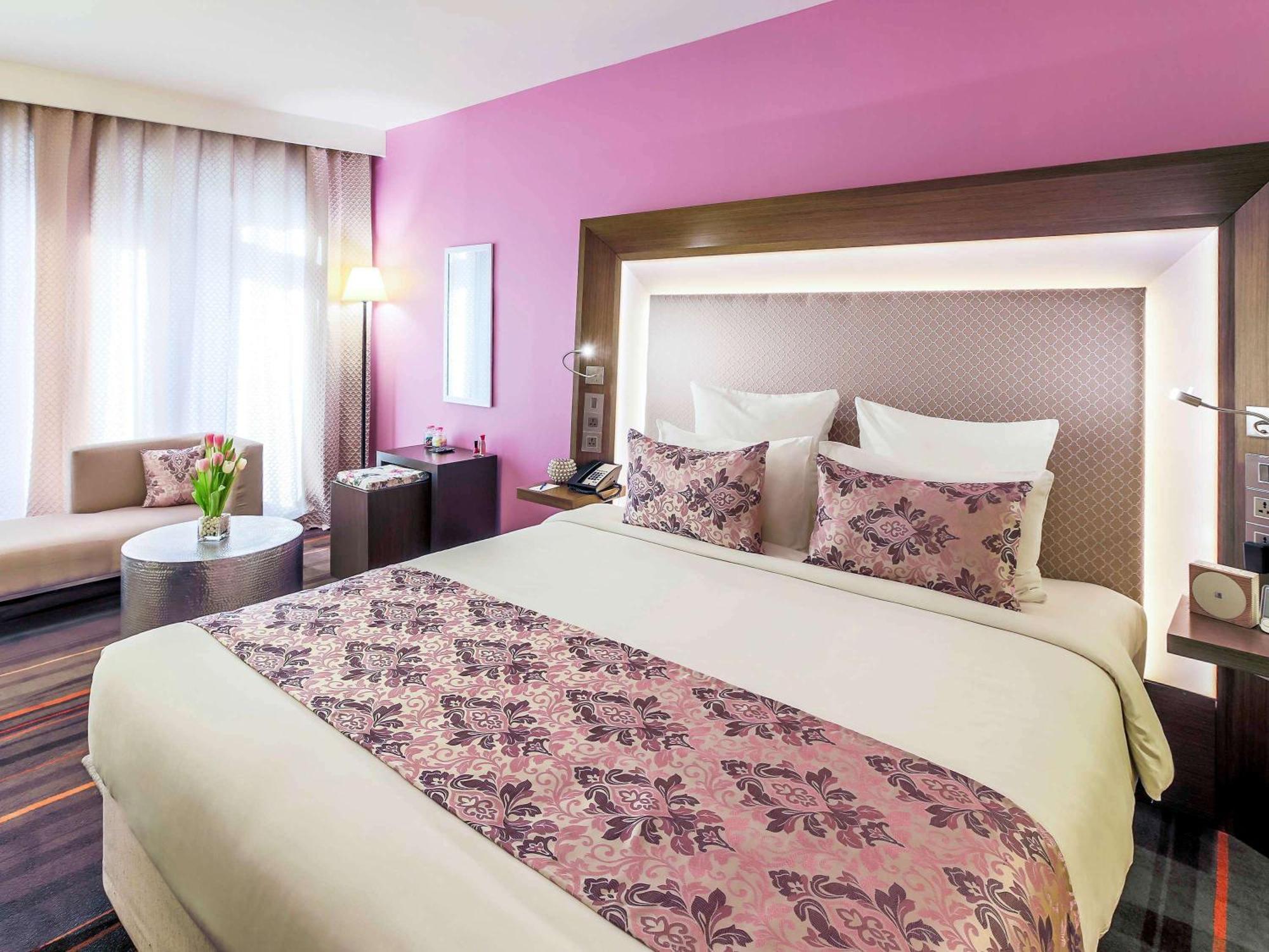 Novotel New Delhi Aerocity Екстериор снимка The photo shows a modern hotel room featuring a queen-sized bed with decorative pillows. The bedding is light-colored with a patterned throw blanket that has shades of pink and purple. Behind the bed, there is a padded headboard with soft lighting. 

