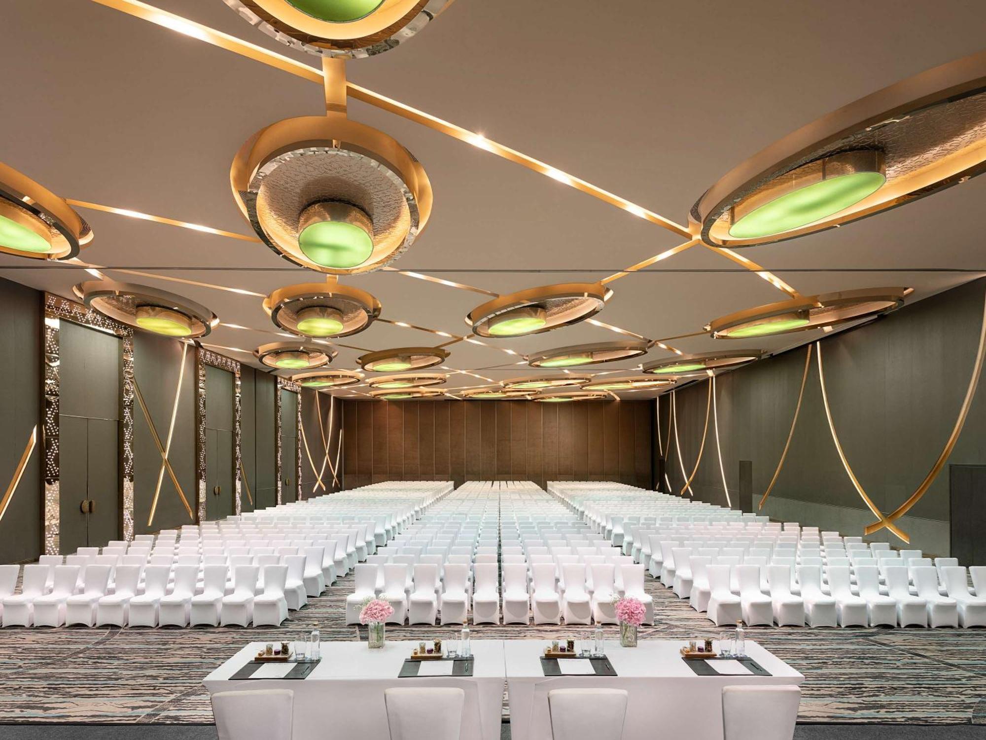 Novotel New Delhi Aerocity Екстериор снимка The photo displays a spacious and elegantly designed event hall. There are rows of white chairs arranged neatly, set up for an audience. At the front, there is a long table, likely for speakers or presenters, adorned with floral arrangements. The cei