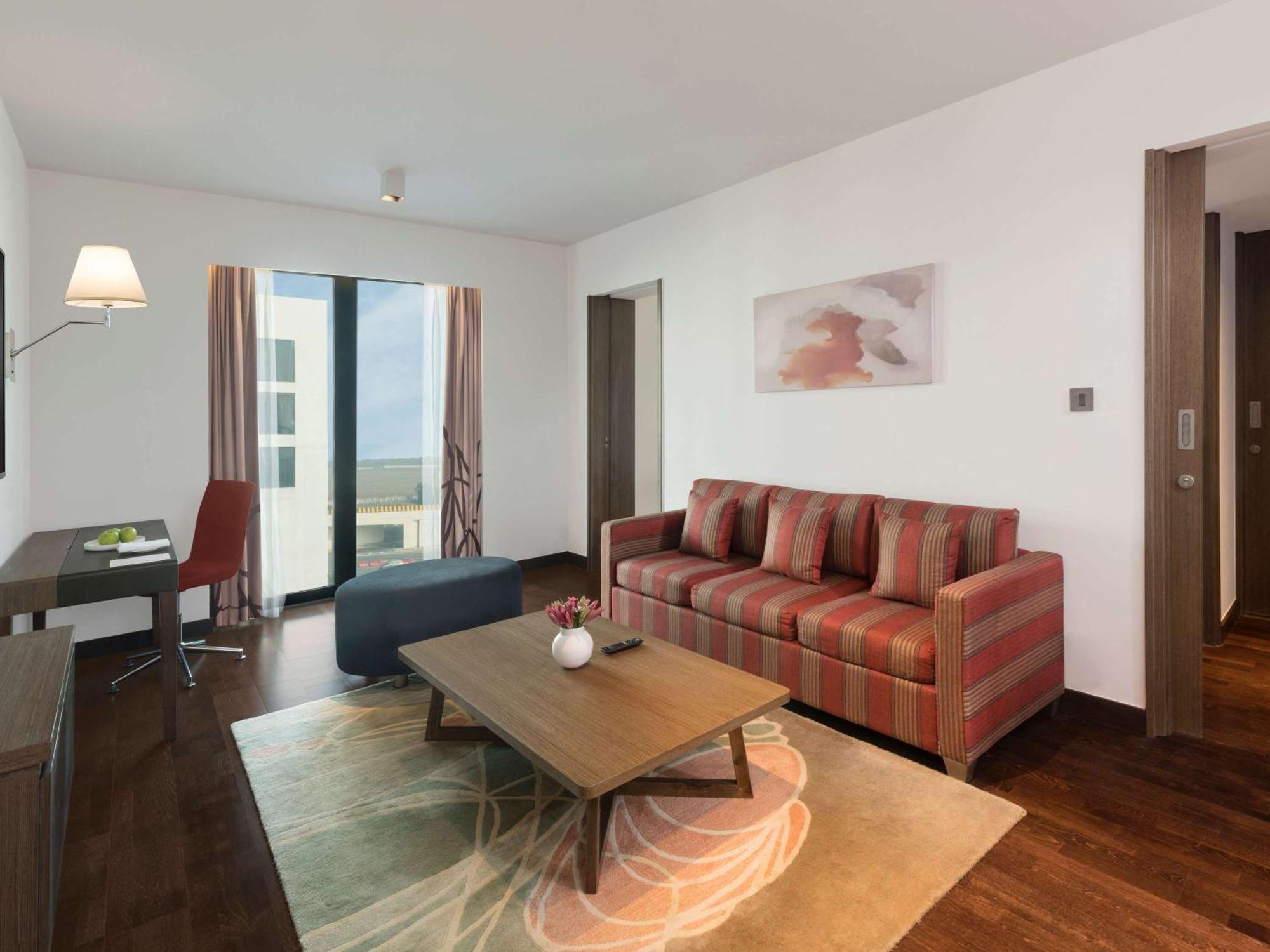 Novotel New Delhi Aerocity Екстериор снимка The photo shows a modern living room area. It features a striped sofa with red and beige tones, arranged facing a coffee table. The coffee table is wooden and has a decorative vase in the center. There's a large area rug underneath, adding texture to