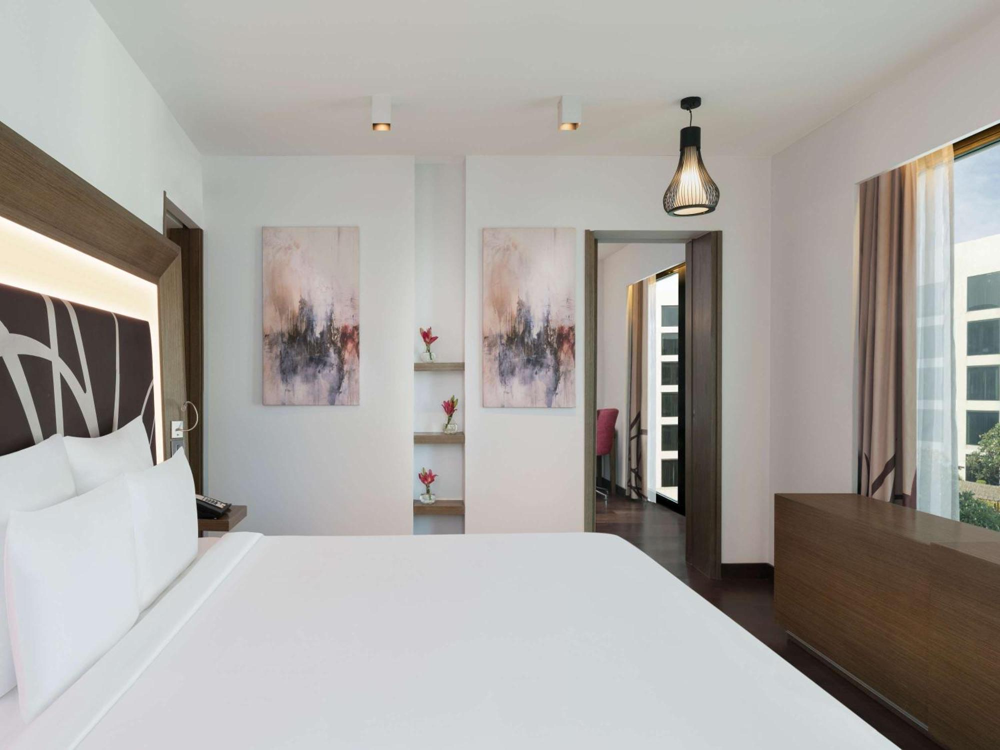 Novotel New Delhi Aerocity Екстериор снимка The photo shows a modern and stylish hotel room. The room features a large bed with a white bedding set, along with a headboard that has a contemporary design. On the walls, there are two abstract paintings that add a touch of color and elegance. To 